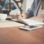 woman writing on a notebook beside teacup and tablet computer